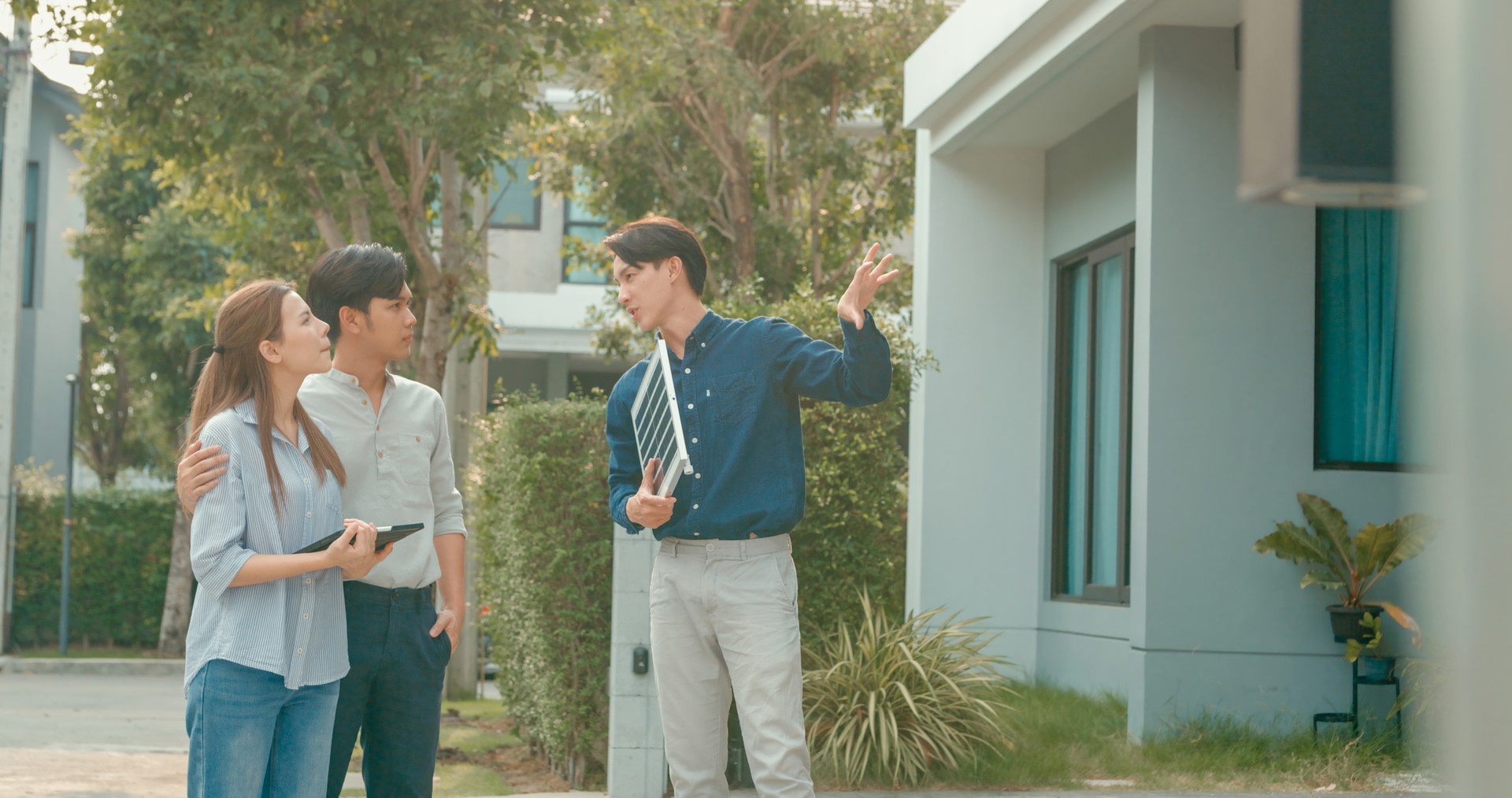 Asian real estate agent demonstrates solar panel benefits to interested young couple outside a modern home. Consulting Eco-Friendly Home with Real Estate Agent.